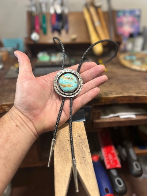 Turquoise bolo tie with stamped silver bezel and braided leather cord, in the maker's hand